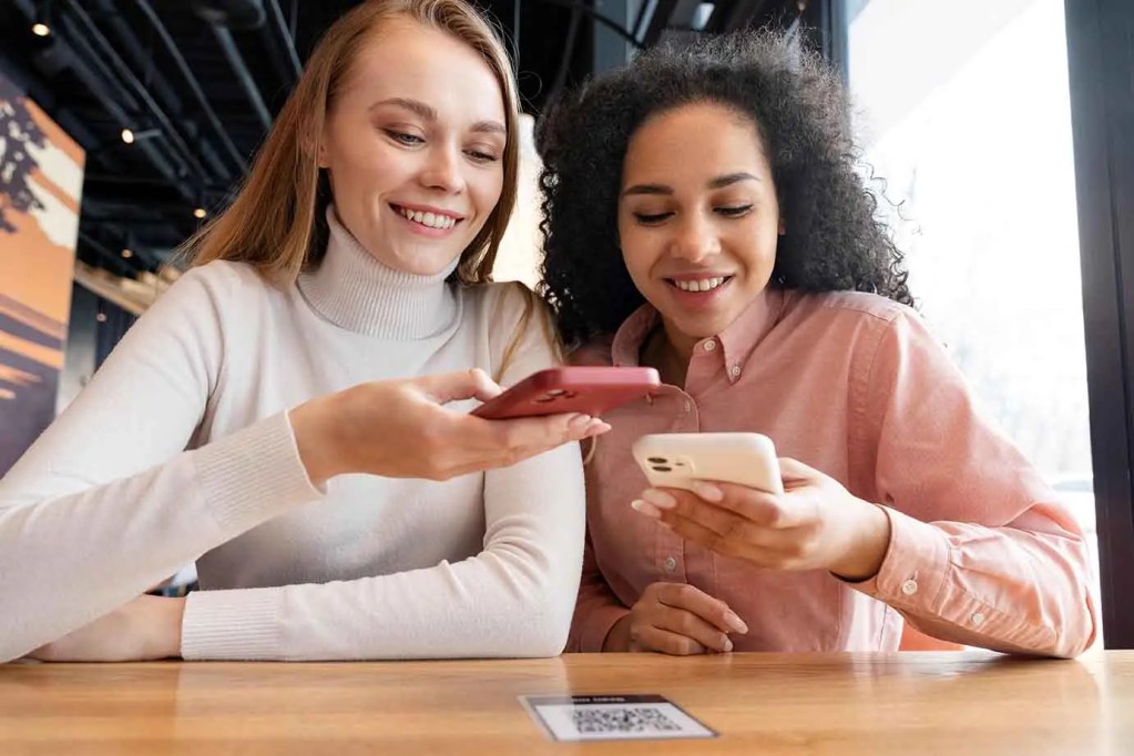 Dos mujeres sentadas en una mesa, usando sus teléfonos móviles para realizar transacciones digitales en un ambiente acogedor.