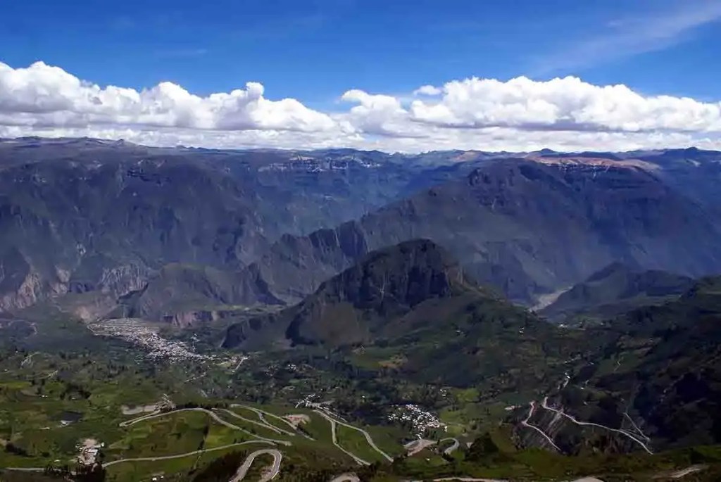 Vista panorámica de montañas y un valle altoandino en Perú, con nubes en el cielo y un asentamiento visible en el fondo.