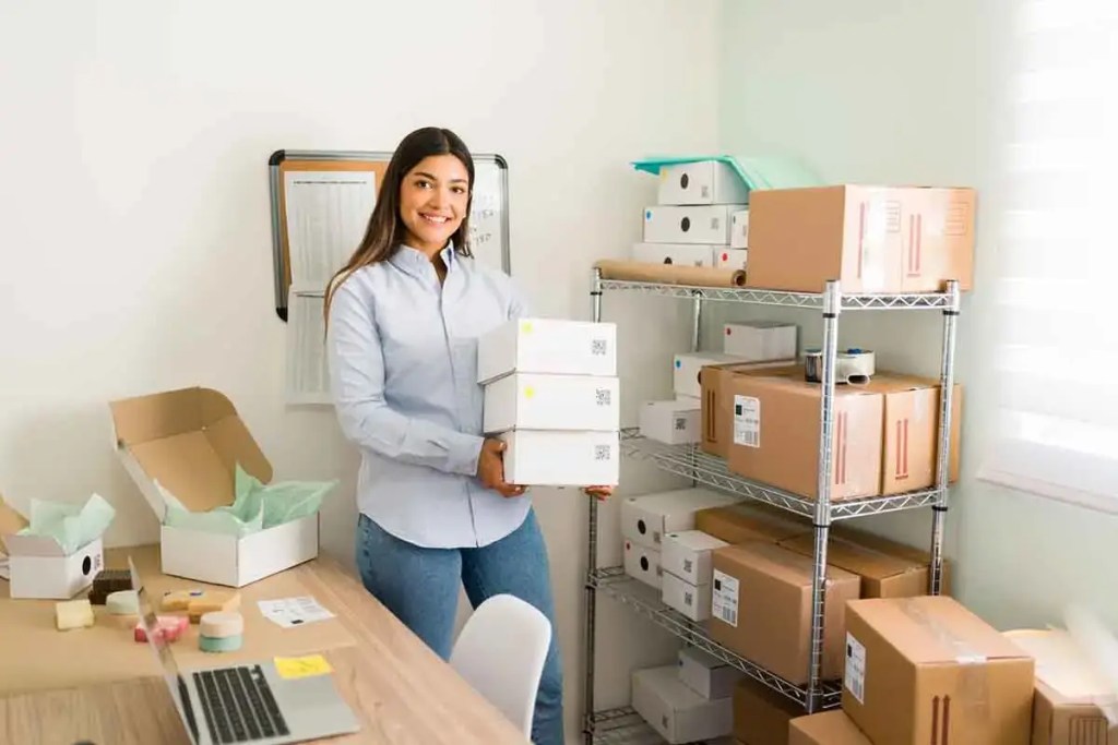 Mujer sonriente sosteniendo cajas de productos en un entorno de oficina de pequeñas empresas, con estanterías llenas de cajas y un escritorio con una computadora portátil.