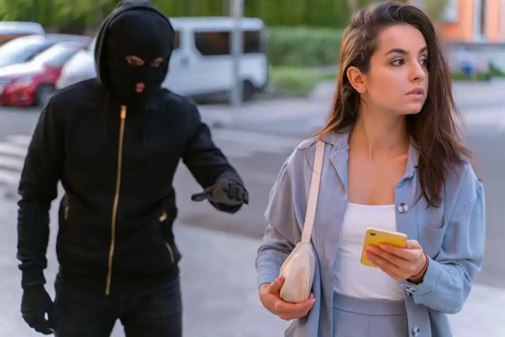 Una mujer aparentemente insegura mira a su alrededor mientras un ladrón con máscara se aproxima desde atrás, mostrando un gesto amenazante.