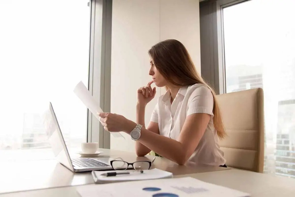 Mujer pensativa sentada en un escritorio, revisando documentos mientras utiliza una computadora portátil, con una taza y gafas sobre la mesa.