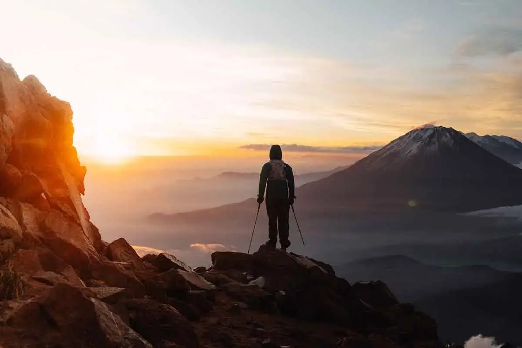 Un montañista de espaldas contempla un amanecer desde la cima de una montaña, con otros volcanes visibles en el horizonte.