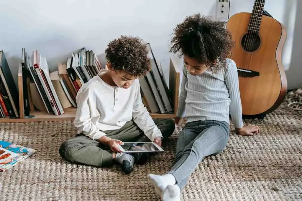 Dos niños sentados en el suelo, mirando una tablet, rodeados de libros y una guitarra en el fondo.