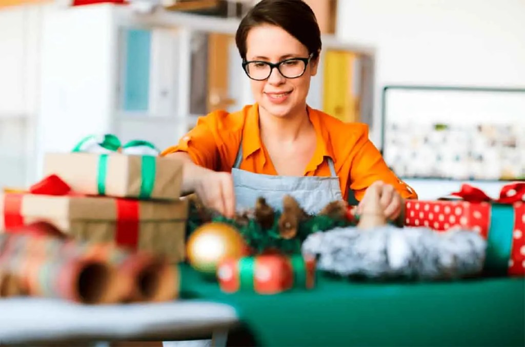 Una mujer sonriente trabaja en la decoración de regalos navideños sobre una mesa, rodeada de cajas envueltas y adornos festivos.