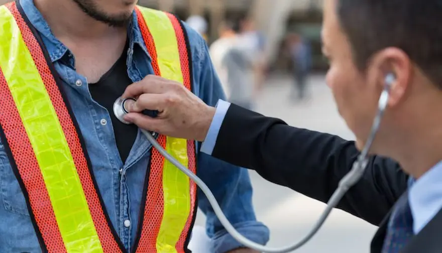 Un médico examina a un trabajador de la construcción que lleva un chaleco reflectante con un estetoscopio.