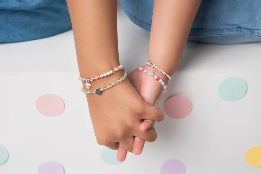 Dos manos de niñas entrelazadas, cada una decorada con pulseras coloridas de la nueva línea de joyería infantil de Bohoo Jewelry, sobre un fondo con círculos de colores.