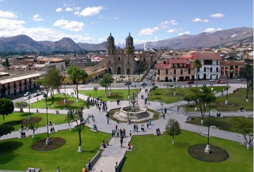 Vista panorámica de una plaza en Cajamarca, Perú, con árboles, personas caminando, edificios coloniales y montañas al fondo.