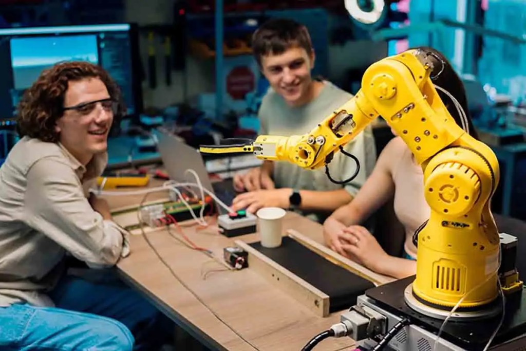 Un grupo de jóvenes observa un robot colaborativo amarillo en un entorno de trabajo, con laptops y equipos electrónicos en la mesa.