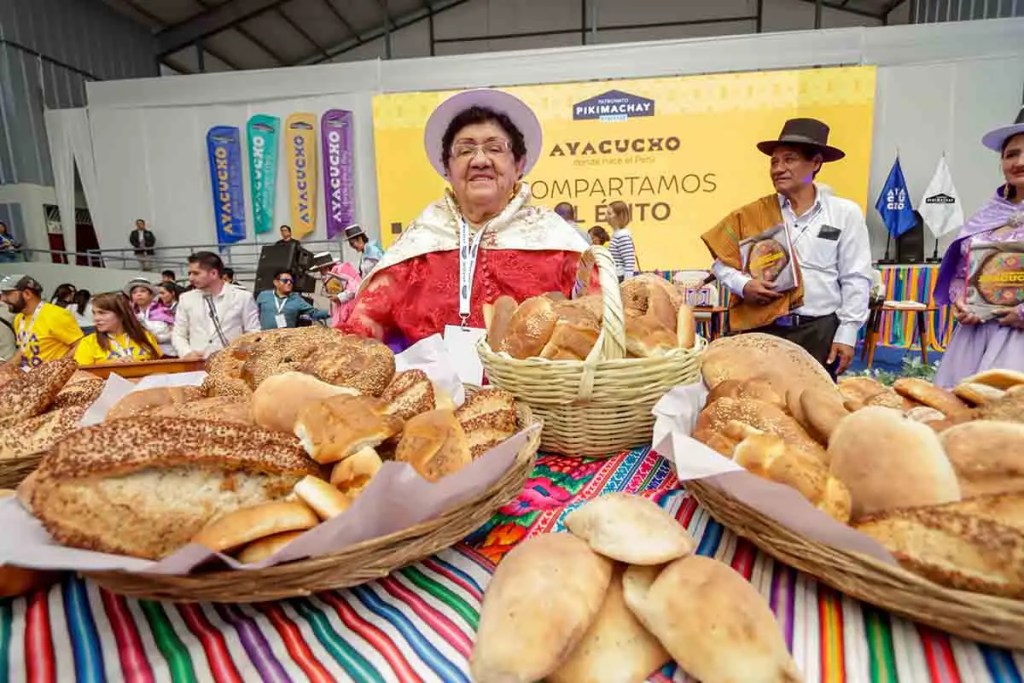 Una mujer mayor vestida con atuendo tradicional sonríe frente a una mesa llena de diferentes tipos de pan ayacuchano en el Foro Gastronómico "Compartamos el Éxito" en Ayacucho, Perú.