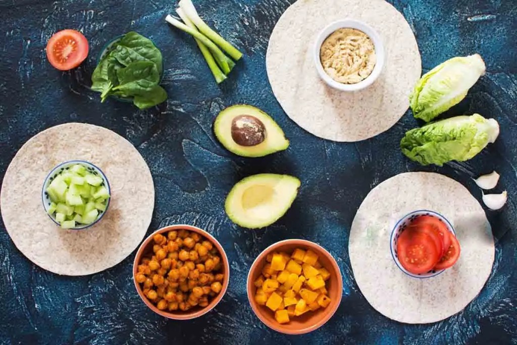 Ingredientes frescos dispuestos sobre una superficie: tortillas, aguacate, espinacas, tomate, pepino, garbanzos y calabaza.