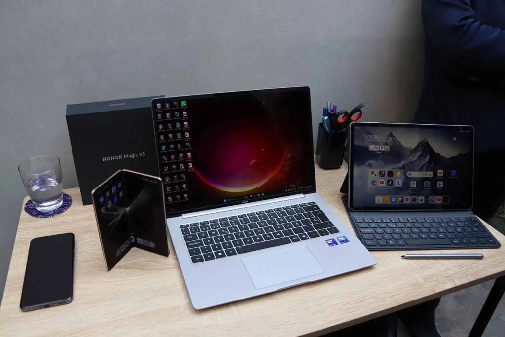 Mesa con dispositivos HONOR: una laptop HONOR MagicBook V5, un teléfono inteligente, una tablet HONOR y un dispositivo plegable, con un vaso de agua y un portalápices al fondo.