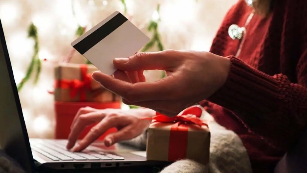 Persona sosteniendo una tarjeta de pago mientras utiliza una computadora portátil, rodeada de regalos envueltos con cintas rojas en un ambiente navideño.