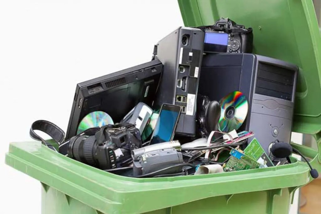 Un contenedor verde lleno de residuos electrónicos, incluyendo computadoras, cámaras, teléfonos móviles y otros dispositivos descartados.