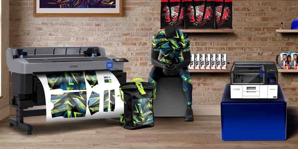 Una impresión de gran formato Epson en un entorno de trabajo, con un modelo humano sentado que lleva ropa personalizada, rodeado de diversos productos impresos y equipos de impresión.