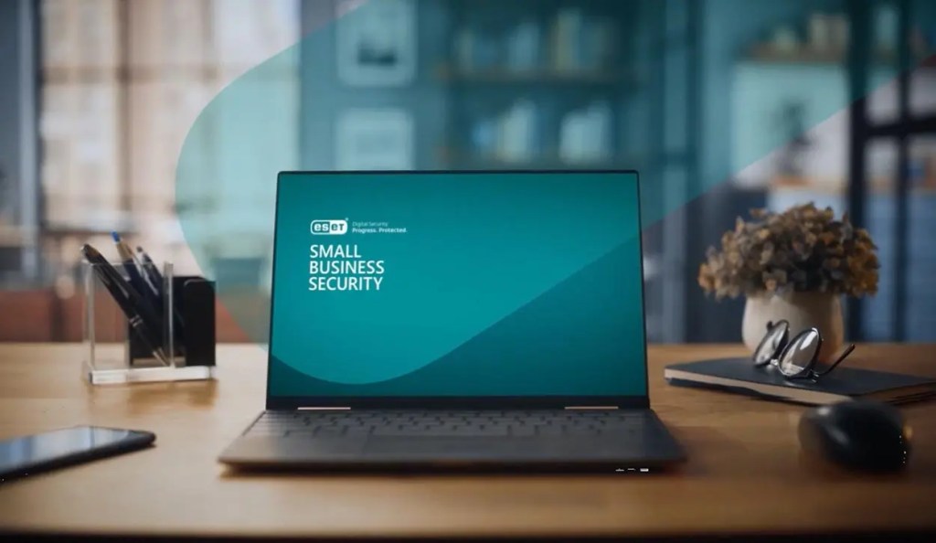 Laptop displaying the ESET Small Business Security logo on a wooden desk, surrounded by a pen holder, glasses, a phone, and a potted plant.