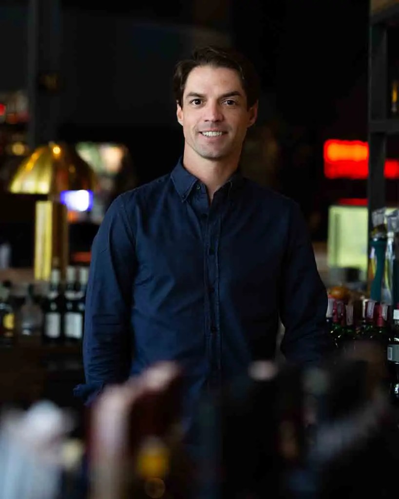 Retrato de un hombre sonriendo, vestido con camisa azul, de pie en un bar con botellas de bebidas espirituosas en el fondo.