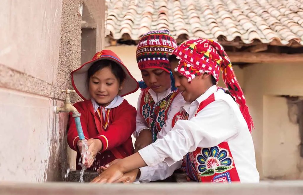 Tres niños vestidos con trajes tradicionales peruanos lavándose las manos en un grifo al aire libre.
