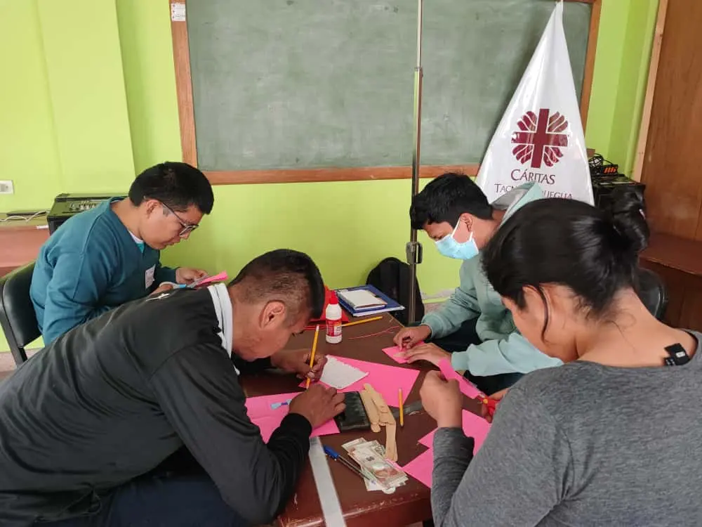 Grupo de jóvenes con discapacidad trabajando en proyectos creativos en una mesa. Al fondo, se observa una pizarra y la bandera de Cáritas.