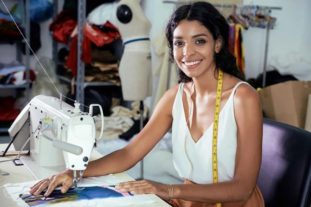Una mujer sonriente sentada frente a una máquina de coser, trabajando en un proyecto de costura en un taller lleno de tela y suministros.