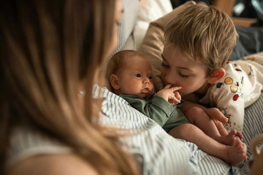 Un niño pequeño acaricia la mano de un bebé en la cama, mientras una madre se asoma suavemente en la imagen, representando un momento tierno entre hermanos.