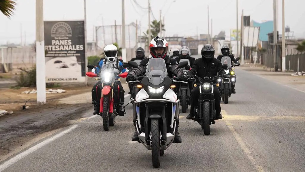 Grupo de motociclistas aventureros en una carretera, liderados por una moto con un piloto en casco, mientras pasan por un letrero que indica 'PANES ARTESANALES'.