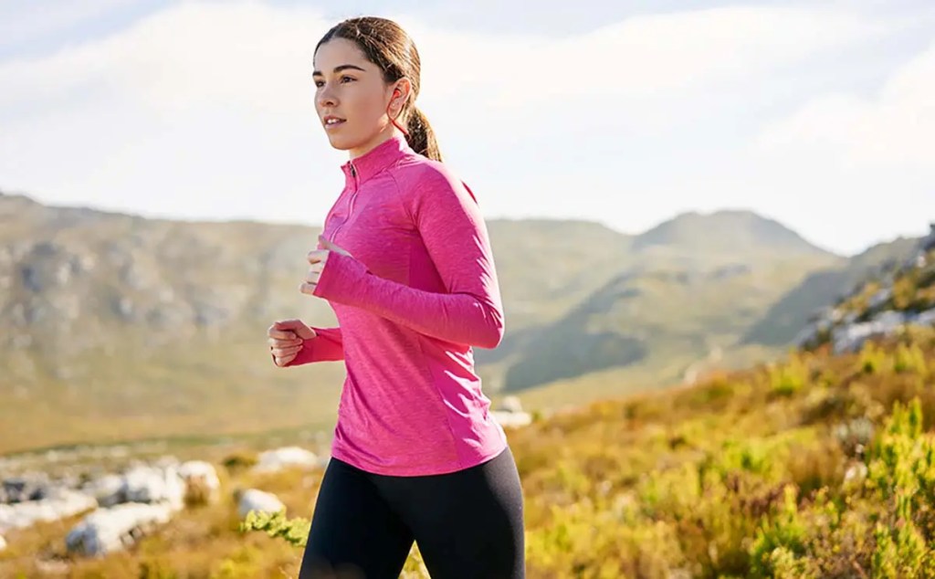 Una persona corriendo en un entorno natural, vistiendo una camiseta de manga larga rosa y pantalones negros, con paisaje montañoso y vegetación al fondo.