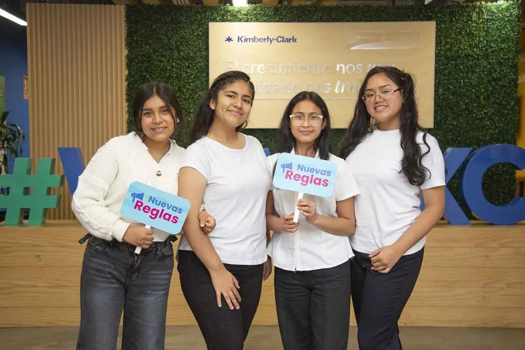 Cuatro adolescentes posando juntas, sosteniendo letreros de 'Nuevas Reglas' en un evento, con un fondo decorado con elementos de Kimberly-Clark.