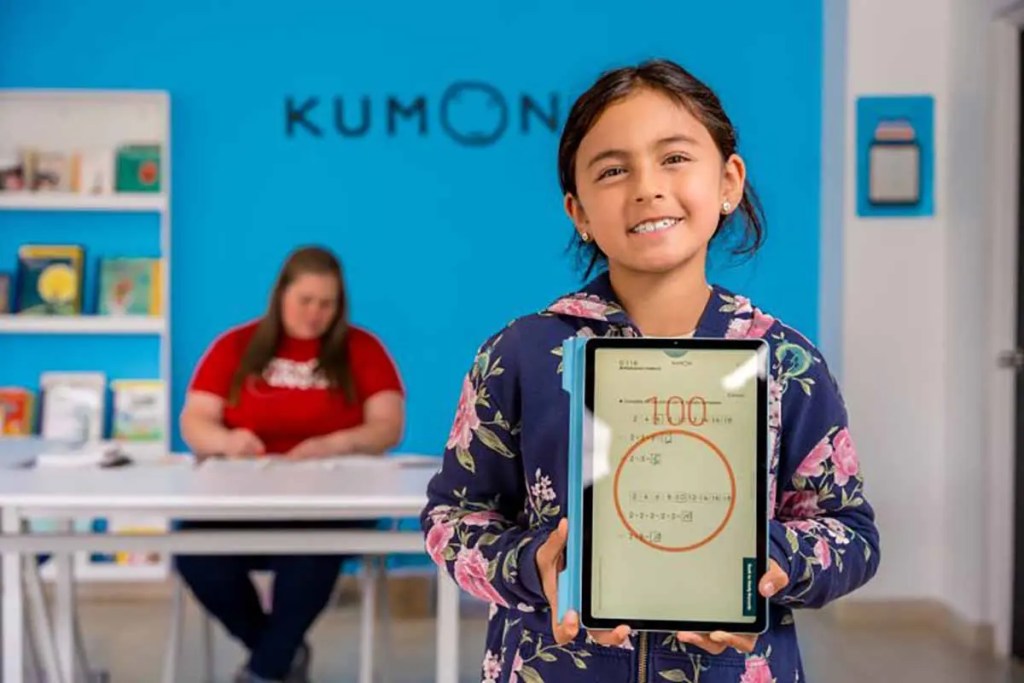 Niña sonriente sosteniendo una tablet con un resultado de 100, en un aula de Kumon con fondo azul y estantería de libros.