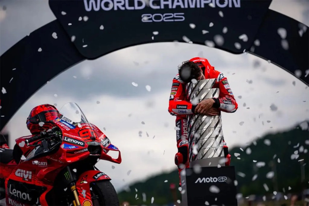 Un piloto de moto vestido con un traje de Ducati celebra la victoria abrazando un trofeo bajo un arco que dice 'World Champion 2025', con confeti cayendo alrededor.