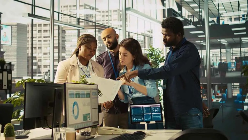 Un grupo de cuatro profesionales discute documentos en una oficina moderna. Dos mujeres y dos hombres están enfocados en el trabajo colaborativo, con computadoras y pantallas en el fondo.