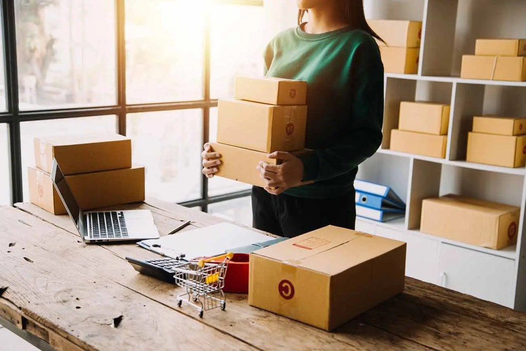 Una persona sostiene varias cajas de cartón en un espacio de trabajo con un ordenador portátil y una mesa de madera, con estanterías llenas de más cajas en el fondo.