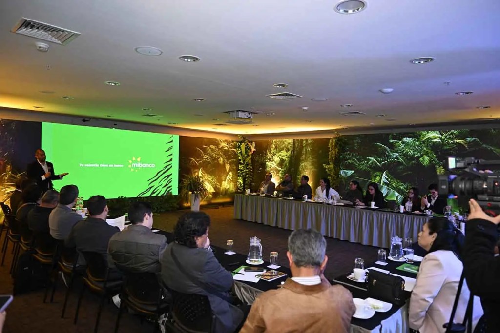 Evento de presentación de Mibanco con una pantalla verde y asistentes sentados en mesas con documentos y bebidas.