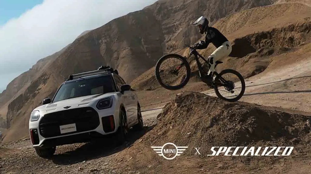 Un ciclista realiza un salto en una bicicleta de montaña cerca de un vehículo MINI, en un entorno montañoso y desértico.