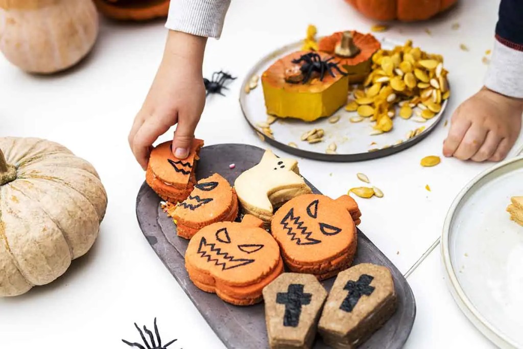 Un niño agarrando galletas decoradas de Halloween en un plato, rodeado de calabazas y semillas de calabaza.