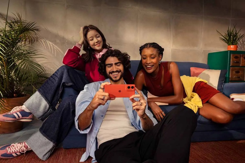 Tres personas sonríen mientras utilizan un smartphone en un ambiente acogedor, sentados en un sofá con plantas decorativas y colores vibrantes de fondo.