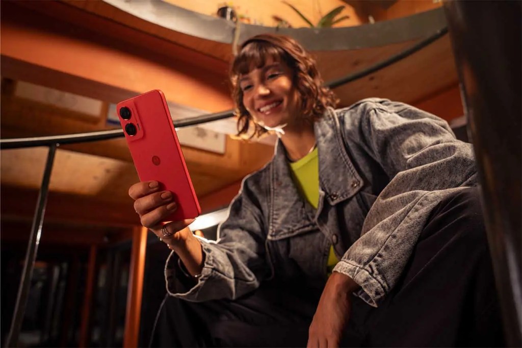 Una persona sonriendo mientras sostiene un smartphone rojo, sentada en una escalera con un entorno moderno.