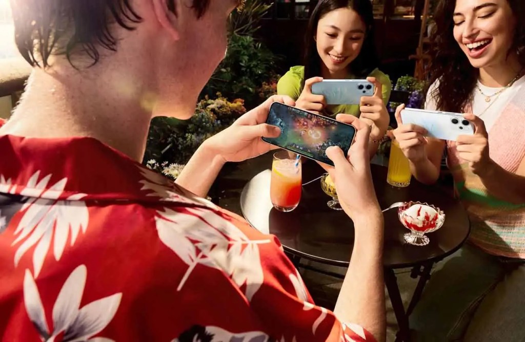 Un grupo de amigos disfruta de una sesión de gaming móvil al aire libre, con un foco en un teléfono inteligente en las manos de una persona y otros dos teléfonos visibles entre los demás.