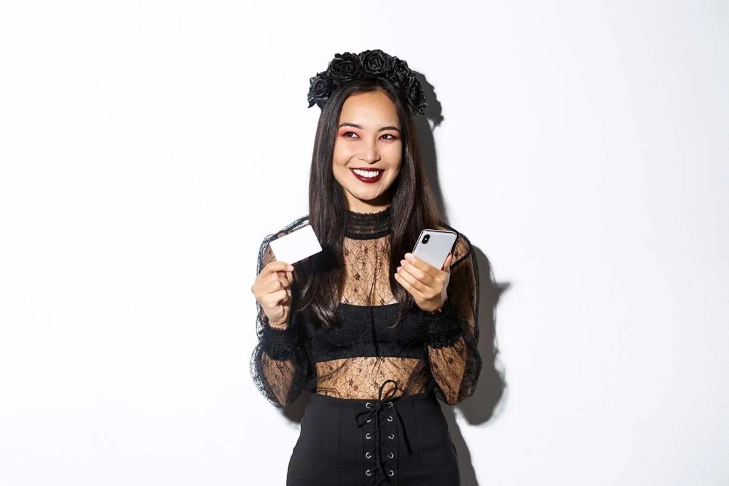 Mujer sonriente con cabello largo y negro, usando una blusa de encaje negro y una corona de flores oscuras, sostiene un teléfono móvil en una mano y una tarjeta en la otra, posando frente a un fondo blanco.