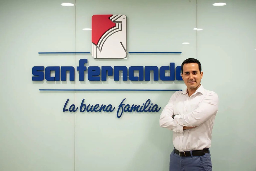 Imagen del logo de San Fernando, una empresa avícola en Perú, acompañado por un hombre vestido con camisa blanca que se muestra sonriente y con los brazos cruzados, en un entorno de oficina.
