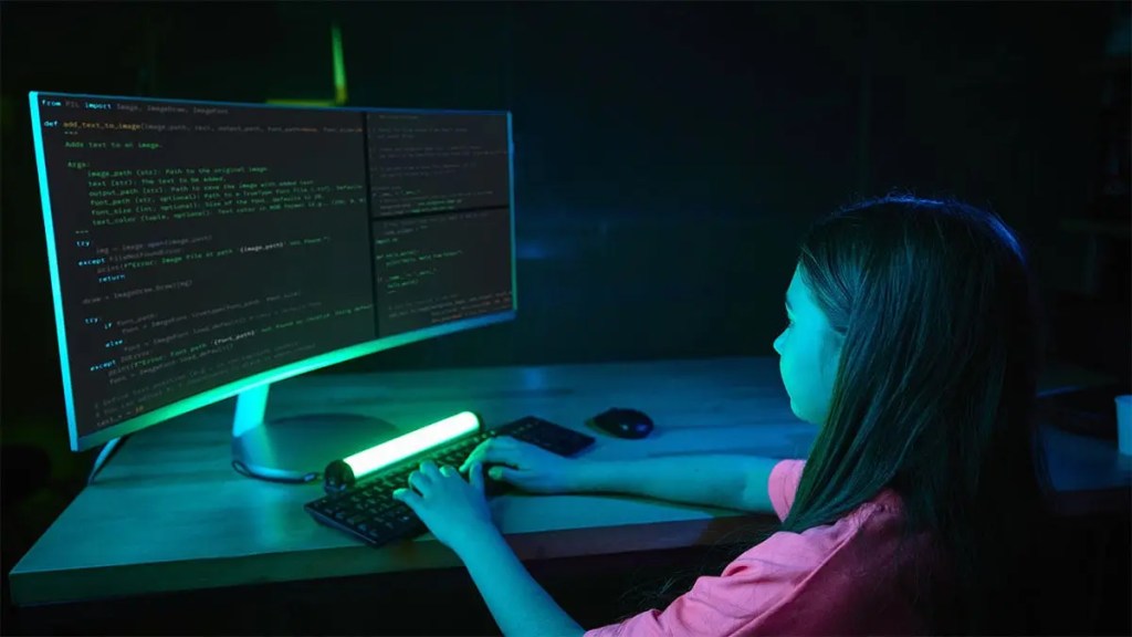 Niña usando un ordenador con código en la pantalla, iluminada por luces en un ambiente oscuro.