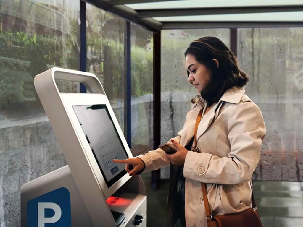 Una mujer interactuando con un monitor táctil en un entorno comercial, utilizando un dispositivo móvil en una mano mientras toca la pantalla.