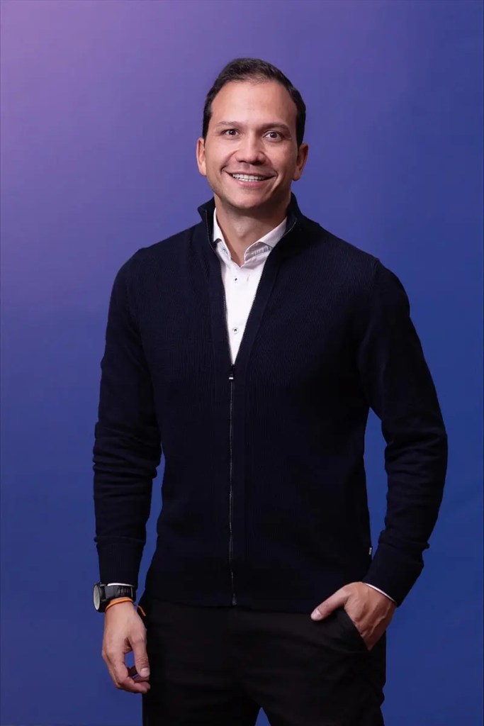 Retrato de Carlos Tovar, nuevo Vicepresidente de Productos para la Región Andina de Visa, sonriendo y luciendo un suéter oscuro frente a un fondo azul.