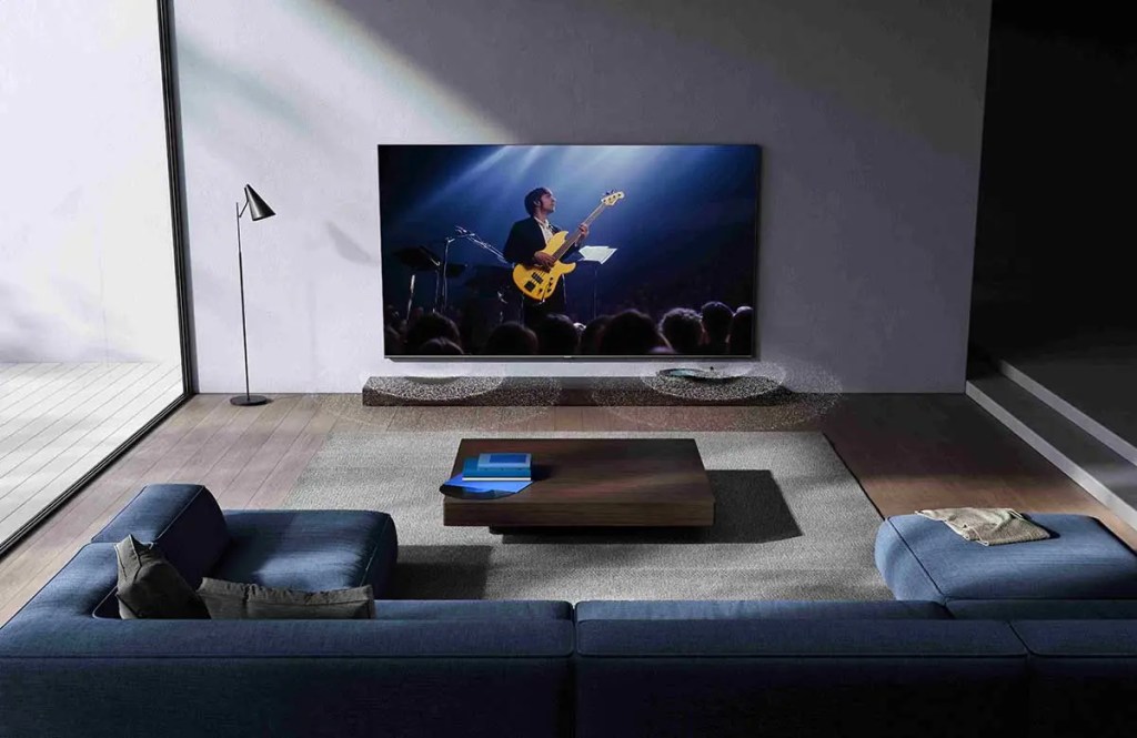 Sala moderna con un televisor Xiaomi mostrando a un músico en vivo, acompañado de un sofá azul y una lámpara de pie, creando un ambiente elegante y acogedor.
