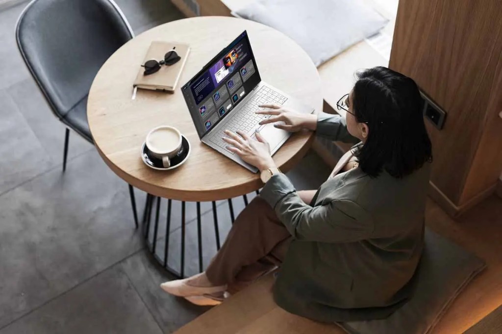 Mujer trabajando en una laptop en una mesa de café, con una taza de café y unas gafas de sol sobre la mesa.