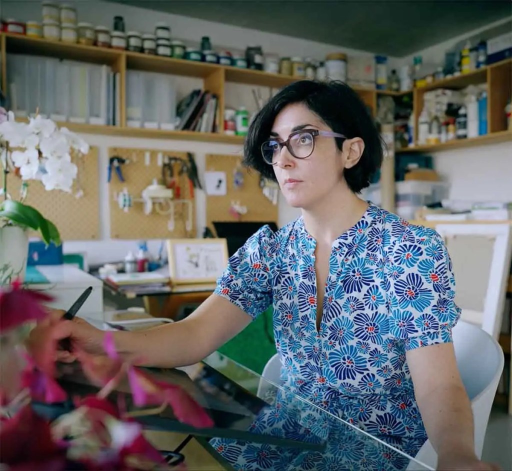 Una mujer con gafas y cabello oscuro trabaja en un escritorio, utilizando una tableta gráfica en un entorno creativo con estanterías de materiales y plantas ornamentales en el fondo.