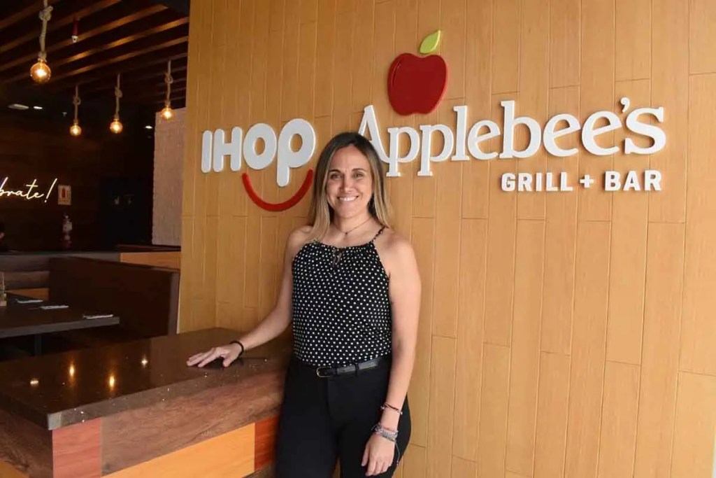 Una mujer posa frente al cartel de IHOP y Applebee's en su nueva ubicación, sonriendo con un fondo moderno y acogedor.