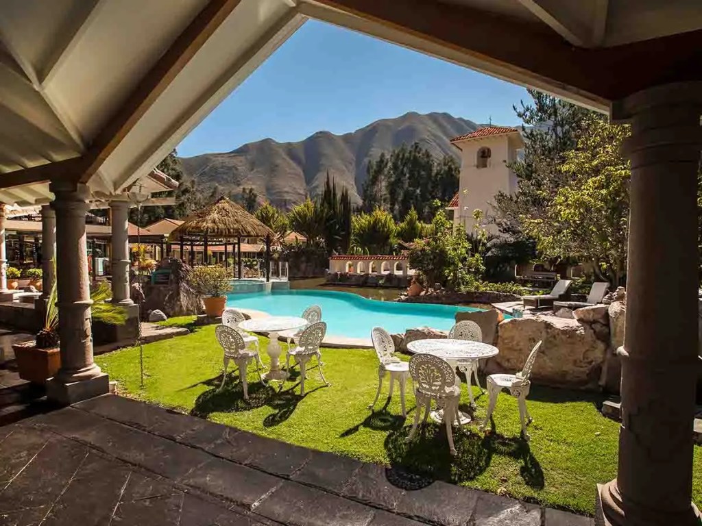 Vista de un área de piscina rodeada de jardines y montañas en un hotel, con mesas y sillas blancas dispuestas en el césped.
