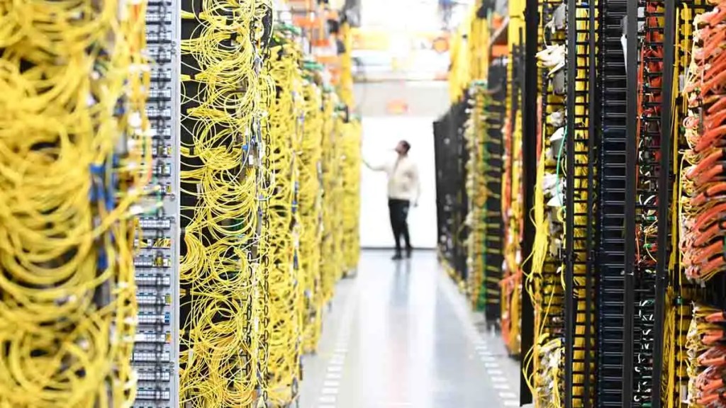 Pasillo de un centro de datos con filas de servidores organizados, cables amarillos y un individuo en el fondo inspeccionando la infraestructura.