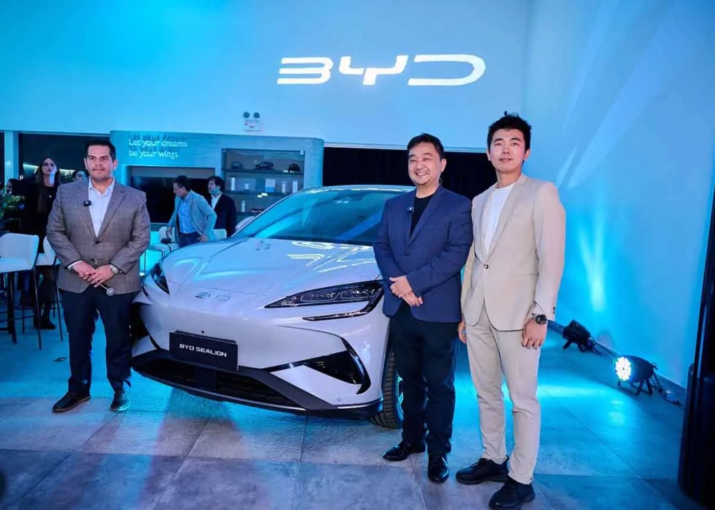 Tres hombres posando frente al nuevo SUV eléctrico BYD Sealion en un evento de lanzamiento, con un fondo iluminado en azul y el logo de BYD proyectado en la pared.