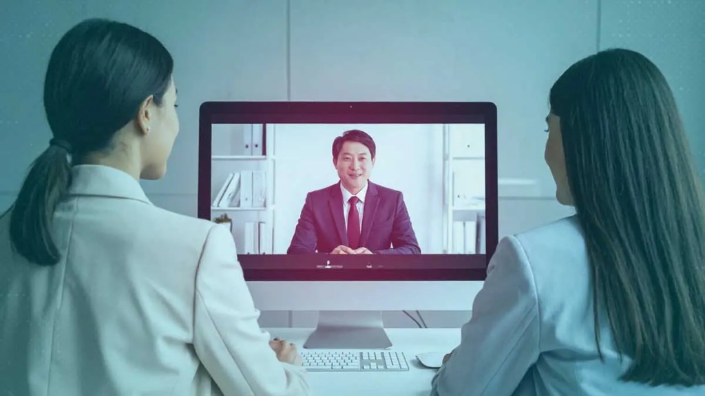 Dos mujeres en trajes de negocio participan en una videoconferencia con un hombre que se presenta en la pantalla de un ordenador.